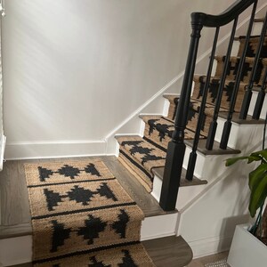 May include: A set of black and tan patterned stair treads with a black wooden handrail. The treads are made of a natural fibre material, possibly jute or sisal.
