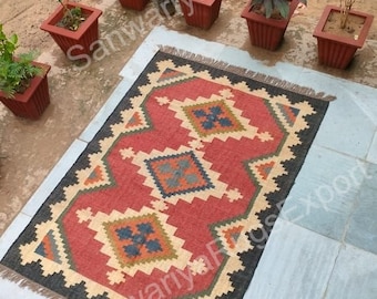 May include: A red, black, and cream colored rug with a geometric pattern. The rug has fringe on all four sides and is laid out on a tiled floor.