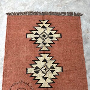 May include: A rust-colored rug with a geometric pattern in black and white. The rug has a fringe border.