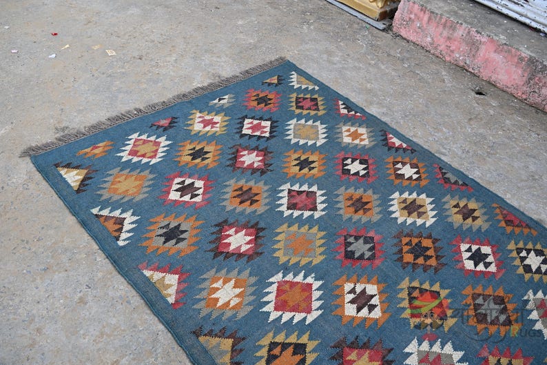 May include: A rectangular blue rug with a repeating geometric pattern of squares and diamonds in shades of orange, red, white, and black. The rug has a fringed edge.