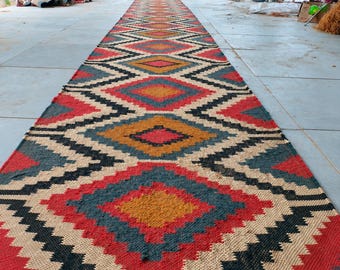 May include: A long, rectangular runner rug featuring a repeating diamond pattern in shades of red, mustard yellow, dark blue, black, and cream. The rug has a woven texture and is finished with a fringe of brown tassels along the bottom edge.