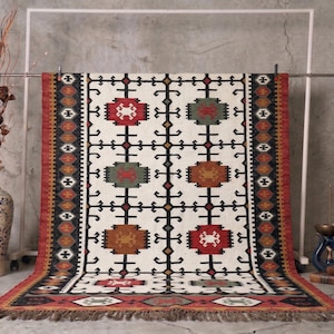 May include: A rectangular, cream-colored rug with a geometric pattern of red, green, and brown shapes. The rug has a fringed edge and is displayed on a metal rack. The rug's design includes black and brown accents.