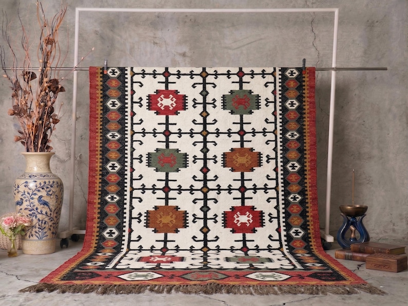 May include: A rectangular, cream-colored rug with a geometric pattern of red, green, and brown shapes. The rug has a fringed edge and is displayed on a metal rack. The rug's design includes black and brown accents.