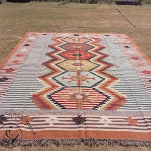Puede incluir: Una alfombra rectangular grande con un patrón geométrico. La alfombra presenta un diseño central en zigzag en tonos de rojo, naranja, amarillo y negro, bordeado por rayas y motivos en forma de estrella. La alfombra está colocada sobre una superficie cubierta de hierba.