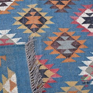 May include: Close-up of a blue woven rug with a geometric pattern. The design features diamond shapes in shades of orange, white, black, and red. The rug has a fringed edge.