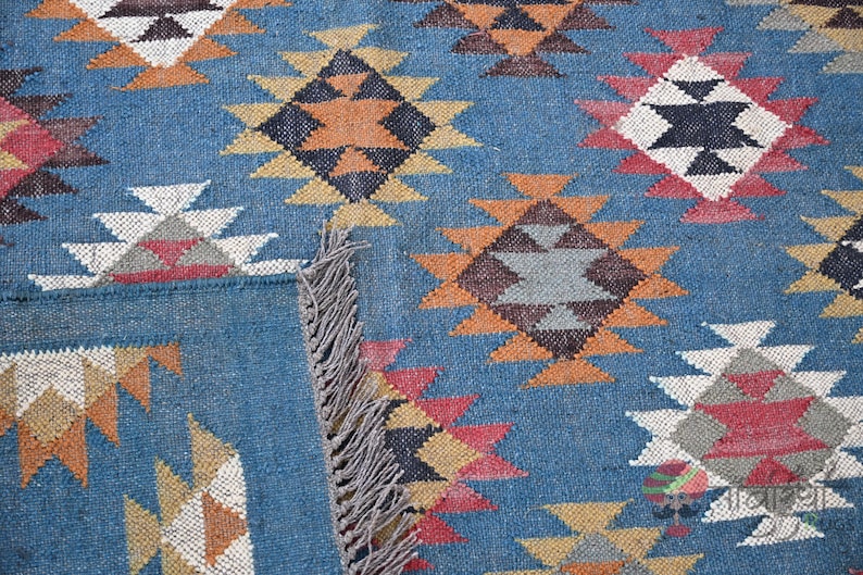 May include: Close-up of a blue woven rug with a geometric pattern. The design features diamond shapes in shades of orange, white, black, and red. The rug has a fringed edge.