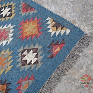 May include: Blue area rug with geometric patterns in orange, red, white, and black. The rug has a fringe on one side. The design features diamond and zig-zag shapes. The rug is made of a woven material.