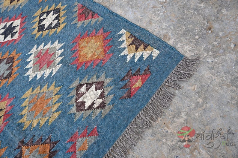 May include: Blue area rug with geometric patterns in orange, red, white, and black. The rug has a fringe on one side. The design features diamond and zig-zag shapes. The rug is made of a woven material.