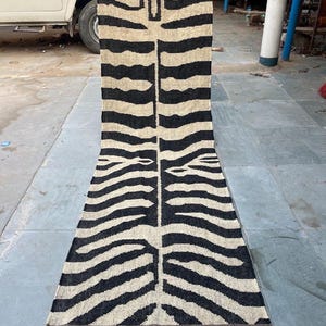 Pode incluir: Um tapete longo e retangular com um design de listras de zebra. O tapete apresenta listras pretas e bege alternadas, com uma borda com franjas. O padrão do tapete se assemelha ao rosto de uma zebra no topo. O tapete é feito de um material tecido.