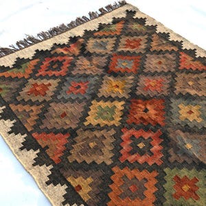 May include: Rectangular rug with a geometric pattern in shades of brown, red, orange, and green. The rug has a beige border and fringe. The design features repeating square and diamond shapes.