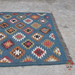 May include: A rectangular blue rug with a geometric diamond pattern in shades of red, orange, white, and brown. The rug has a fringed edge and is placed on a concrete surface. The rug is a decorative home textile.