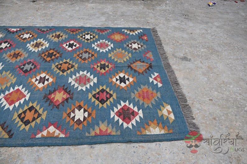 May include: A rectangular blue rug with a geometric diamond pattern in shades of red, orange, white, and brown. The rug has a fringed edge and is placed on a concrete surface. The rug is a decorative home textile.