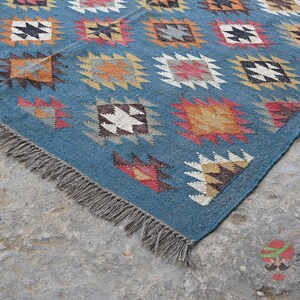 May include: A blue rug with a geometric pattern of squares in various colors, including orange, red, white, and black. The rug has a fringe along one edge and is made of a woven material. The rug is placed on a concrete surface.