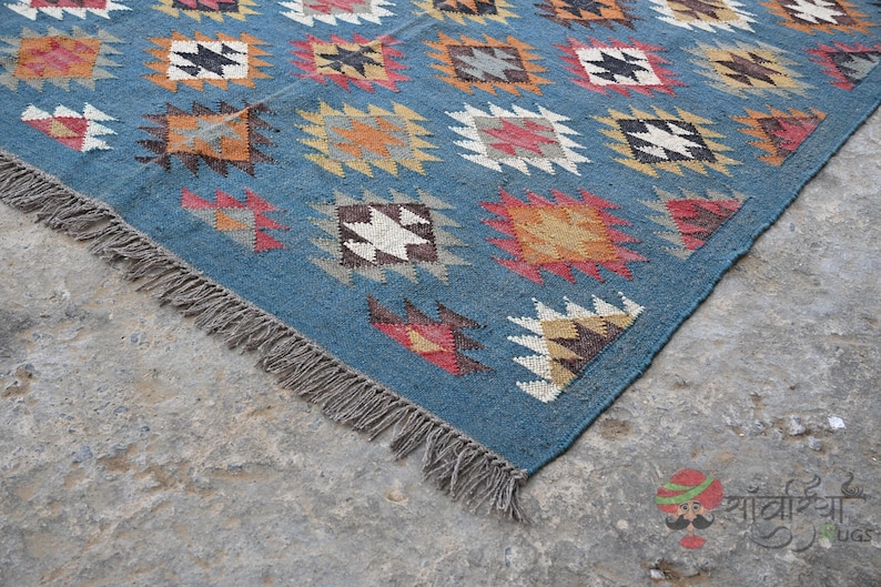 May include: A blue rug with a geometric pattern of squares in various colors, including orange, red, white, and black. The rug has a fringe along one edge and is made of a woven material. The rug is placed on a concrete surface.
