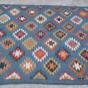 May include: Blue area rug with a repeating diamond pattern in shades of red, orange, black, and white. The rug has a fringe on one end and is made of a woven material.