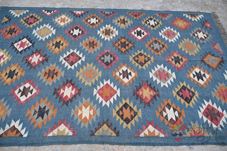 May include: Blue area rug with a repeating diamond pattern in shades of red, orange, black, and white. The rug has a fringe on one end and is made of a woven material.