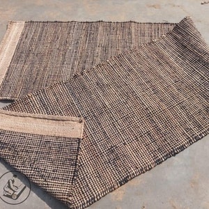May include: A woven rug made of natural fibers in a black and beige color scheme. The rug is folded in half, showing the texture and weave of the material.