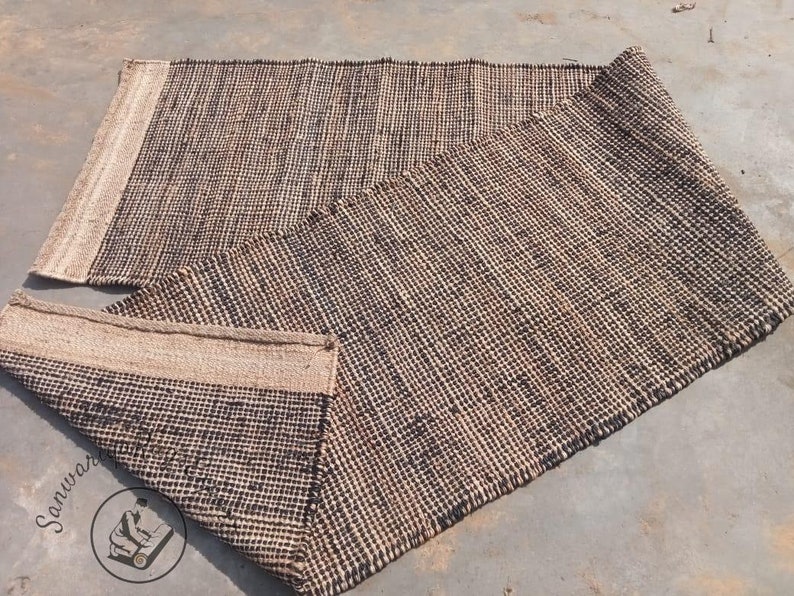 May include: A woven rug made of natural fibers in a black and beige color scheme. The rug is folded in half, showing the texture and weave of the material.