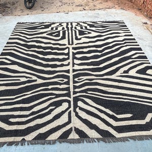 A large black and off-white zebra-patterned rug with fringe lies flat on a concrete surface, with bricks visible in the background.