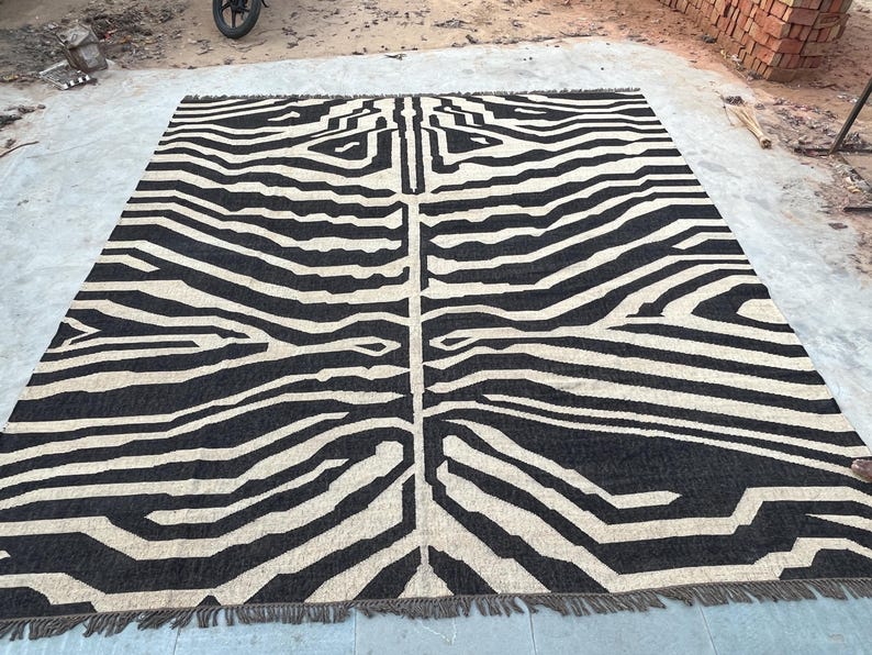 A large black and off-white zebra-patterned rug with fringe lies flat on a concrete surface, with bricks visible in the background.