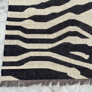 A close-up of a black and off-white zebra-patterned rug with fringe, showing the woven texture on a concrete floor.