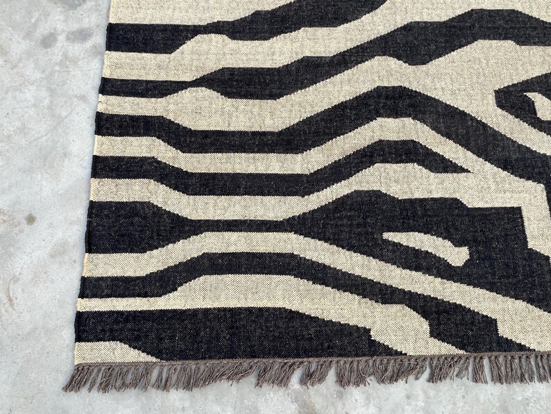 A close-up of a black and off-white zebra-patterned rug with fringe, showing the woven texture on a concrete floor.