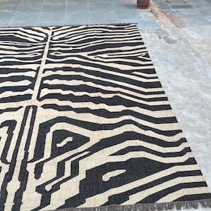 A large black and off-white zebra-patterned rug with fringe lies flat on a concrete floor, with industrial equipment in the background.