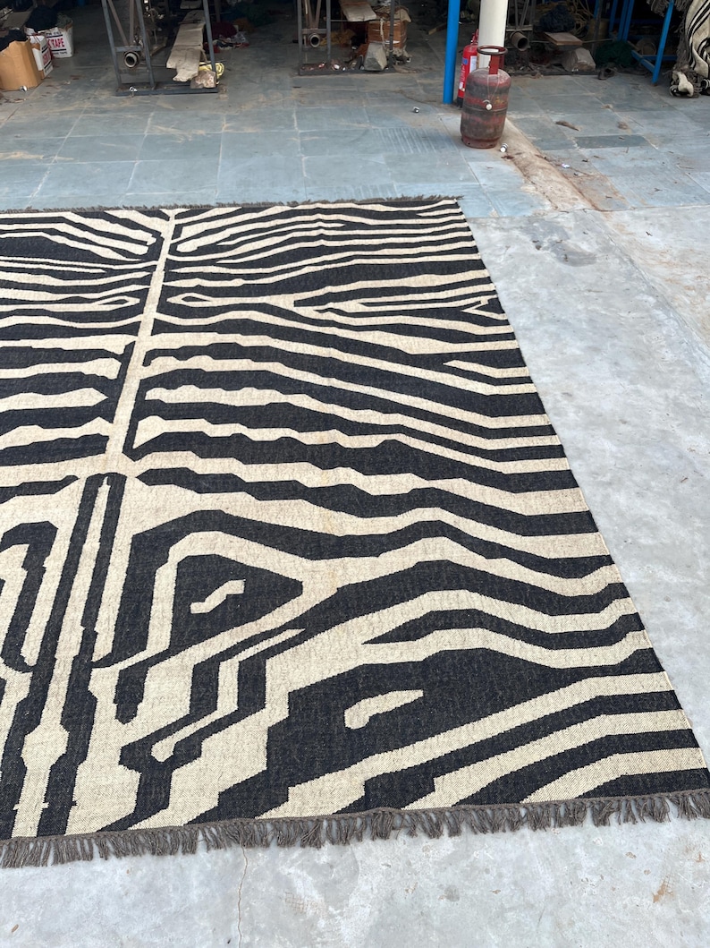 A large black and off-white zebra-patterned rug with fringe lies flat on a concrete floor, with industrial equipment in the background.