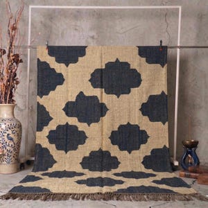 May include: A beige and navy blue patterned rug with a repeating quatrefoil design. The rug has a fringed edge and is displayed on a metal rack. The rug is made of a natural fiber material.
