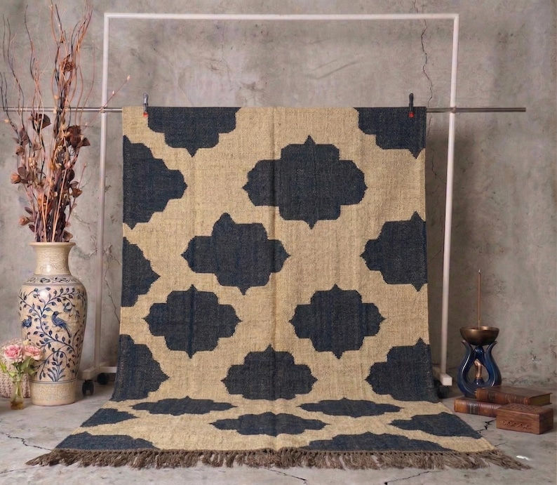May include: A beige and navy blue patterned rug with a repeating quatrefoil design. The rug has a fringed edge and is displayed on a metal rack. The rug is made of a natural fiber material.