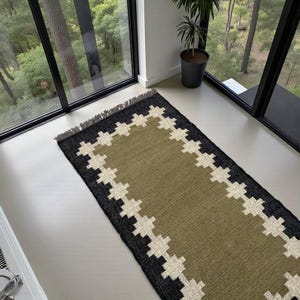 May include: A rectangular area rug featuring a solid olive green centre bordered by a black frame with a repeating white geometric cross pattern. The rug has a short fringe along the top edge and is constructed with a flat woven texture.