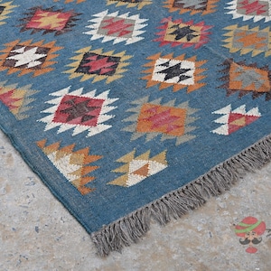 May include: A blue rug with a geometric pattern of colorful diamond shapes. The rug has a fringe on one edge and is made of woven material. The diamond shapes are in shades of red, orange, yellow, and white.