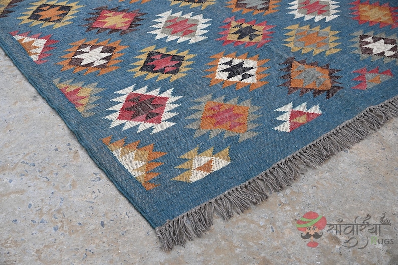 May include: A blue rug with a geometric pattern of colorful diamond shapes. The rug has a fringe on one edge and is made of woven material. The diamond shapes are in shades of red, orange, yellow, and white.