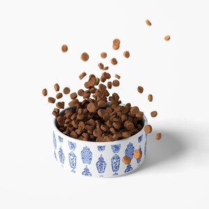 May include: A white ceramic pet bowl with a blue and white Asian-inspired pattern, filled with brown pet food. Some pieces of food are falling from the bowl, creating a dynamic image. The bowl is round and sits on a white surface.