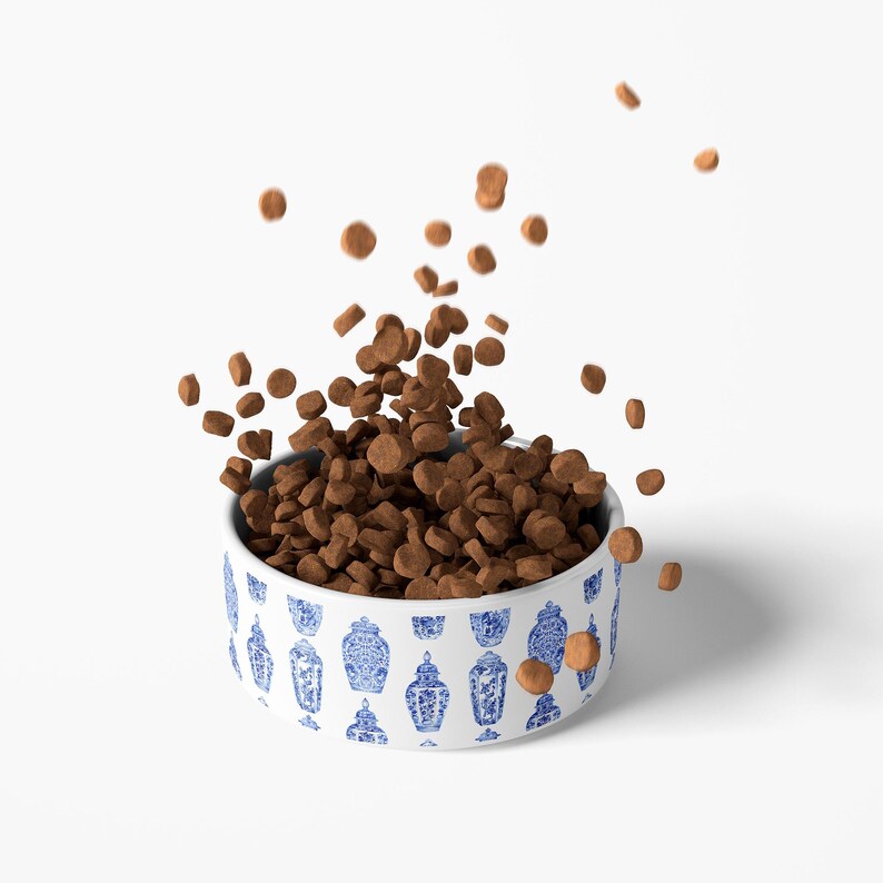 May include: A white ceramic pet bowl with a blue and white Asian-inspired pattern, filled with brown pet food. Some pieces of food are falling from the bowl, creating a dynamic image. The bowl is round and sits on a white surface.