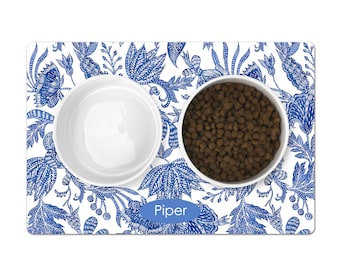 May include: Blue and white patterned pet food mat with two white ceramic bowls. The mat has the name "Piper" written in blue.