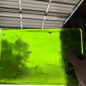 May include: A rectangular piece of bright green glass with a textured surface. The glass is held up, allowing light to pass through. The edges are smooth and rounded. The glass has small air bubbles.