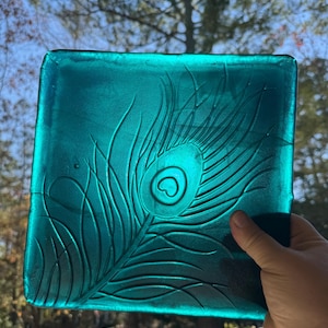 May include: A square, teal-colored glass plate with a peacock feather design. The feather is etched into the glass, creating a textured surface. The plate is held up against a blurred outdoor background.