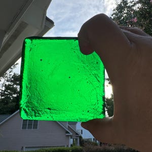 May include: A square, vibrant green glass piece is held up to the sunlight. The glass has a textured surface and appears to be handmade. The bright green color contrasts with the sky and surrounding environment.