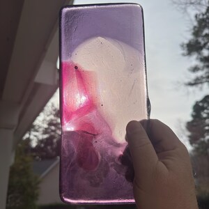 May include: A rectangular, translucent art piece with a blend of colors. The top is a deep purple, transitioning to white and vibrant pink. The piece is held by a hand, with a blurred outdoor background.