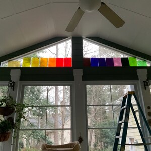 Pode incluir: Uma sala de estar com uma grande janela que apresenta um arco-&iacute;ris de pain&eacute;is de vidro coloridos. A janela tem forma triangular e est&aacute; localizada acima de duas janelas grandes. Uma escada est&aacute; encostada em uma das janelas.