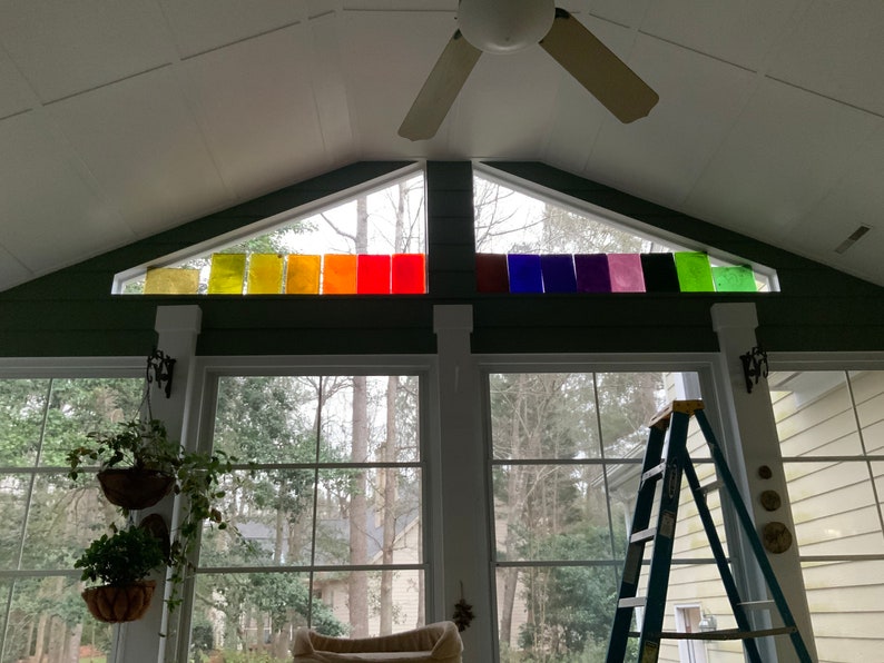 Pode incluir: Uma sala de estar com uma grande janela que apresenta um arco-&iacute;ris de pain&eacute;is de vidro coloridos. A janela tem forma triangular e est&aacute; localizada acima de duas janelas grandes. Uma escada est&aacute; encostada em uma das janelas.