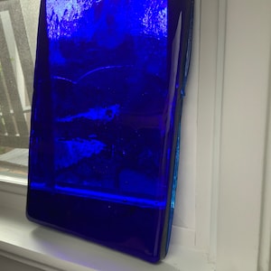 May include: A rectangular, deep blue glass art piece with a textured surface. The artwork is positioned on a white windowsill, with a glimpse of a window and outdoor scenery in the background.