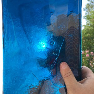 May include: A rectangular, transparent, cobalt blue glass object. The glass has a textured surface with small bubbles and a long, thin crack. The object is held by a hand, with a bright light reflecting off the glass.