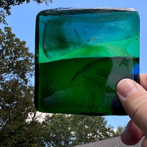 May include: A square piece of green and blue glass with a layered effect. The glass is translucent and has a slight ripple pattern.