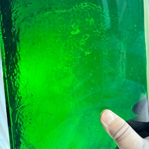 May include: Close-up of a rectangular, transparent, emerald green glass object. The glass has a textured surface with small bubbles and imperfections. A finger is pointing at the glass.