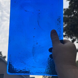 May include: A rectangular, transparent, vibrant blue glass piece is held up against a cloudy sky. The glass has a textured surface with small imperfections, and the edges are slightly rounded. The light shines through the glass, highlighting its color and form.