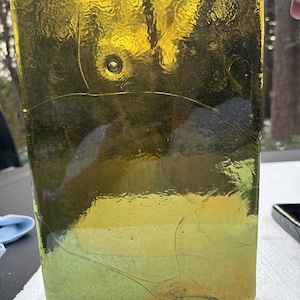 May include: A rectangular, amber-colored glass vase with a textured surface. The vase has a bubble-like detail and visible cracks. The glass appears to be hand-blown, with variations in color and texture. The vase is suitable for home decor.