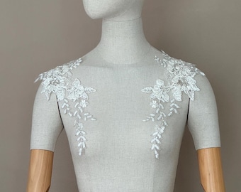 May include: A white lace applique featuring floral and leaf patterns is shown on a mannequin. The applique is designed for the shoulders and upper chest. The mannequin has wooden arms and a neutral-toned torso.