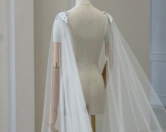 May include: A white tulle bridal cape with delicate lace appliques on the shoulders. The cape is draped over a dress form and has a flowing, ethereal look.